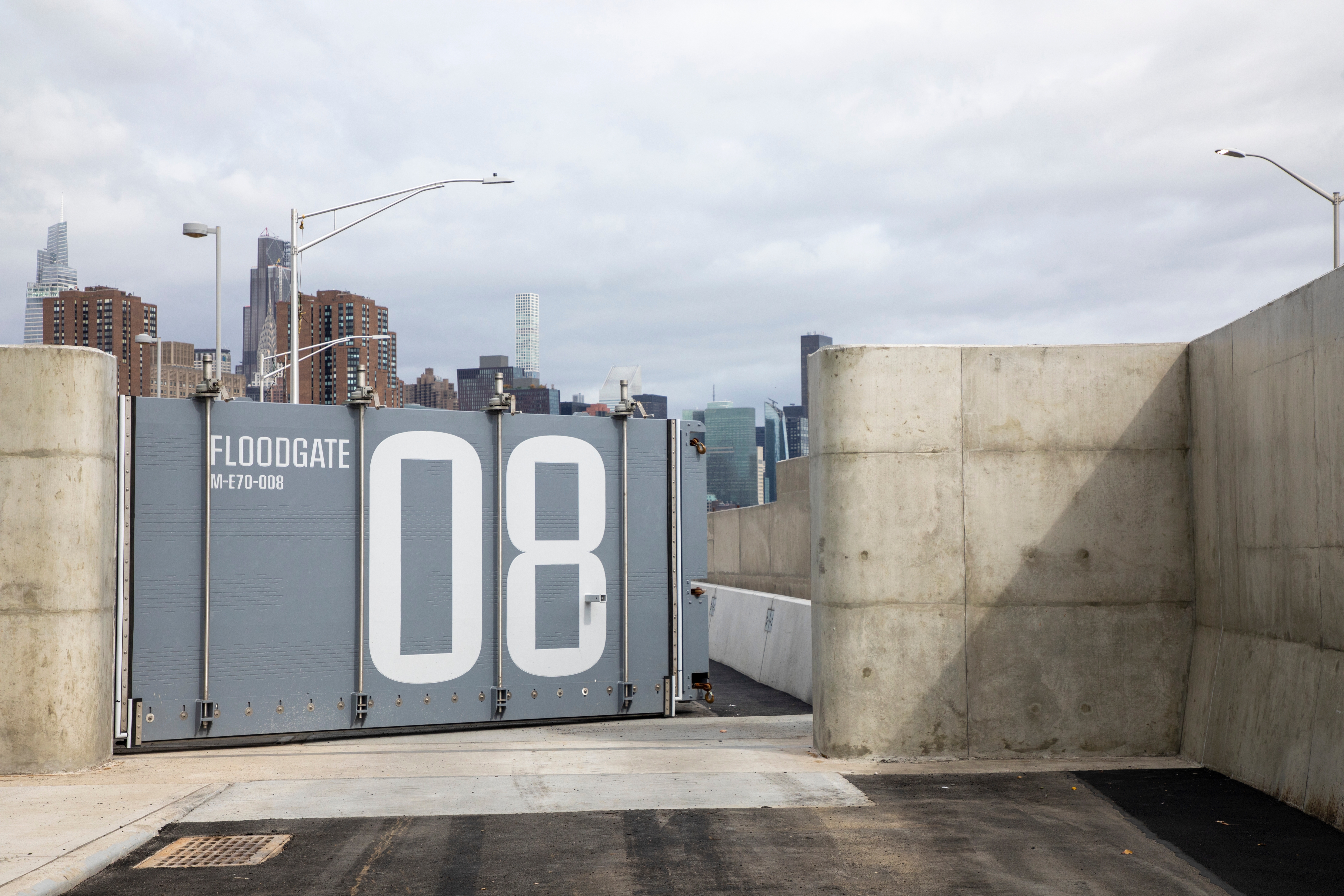 Large gray floodgate labeled "08" blocks a concrete roadway, with a city skyline visible in the background under a cloudy sky.