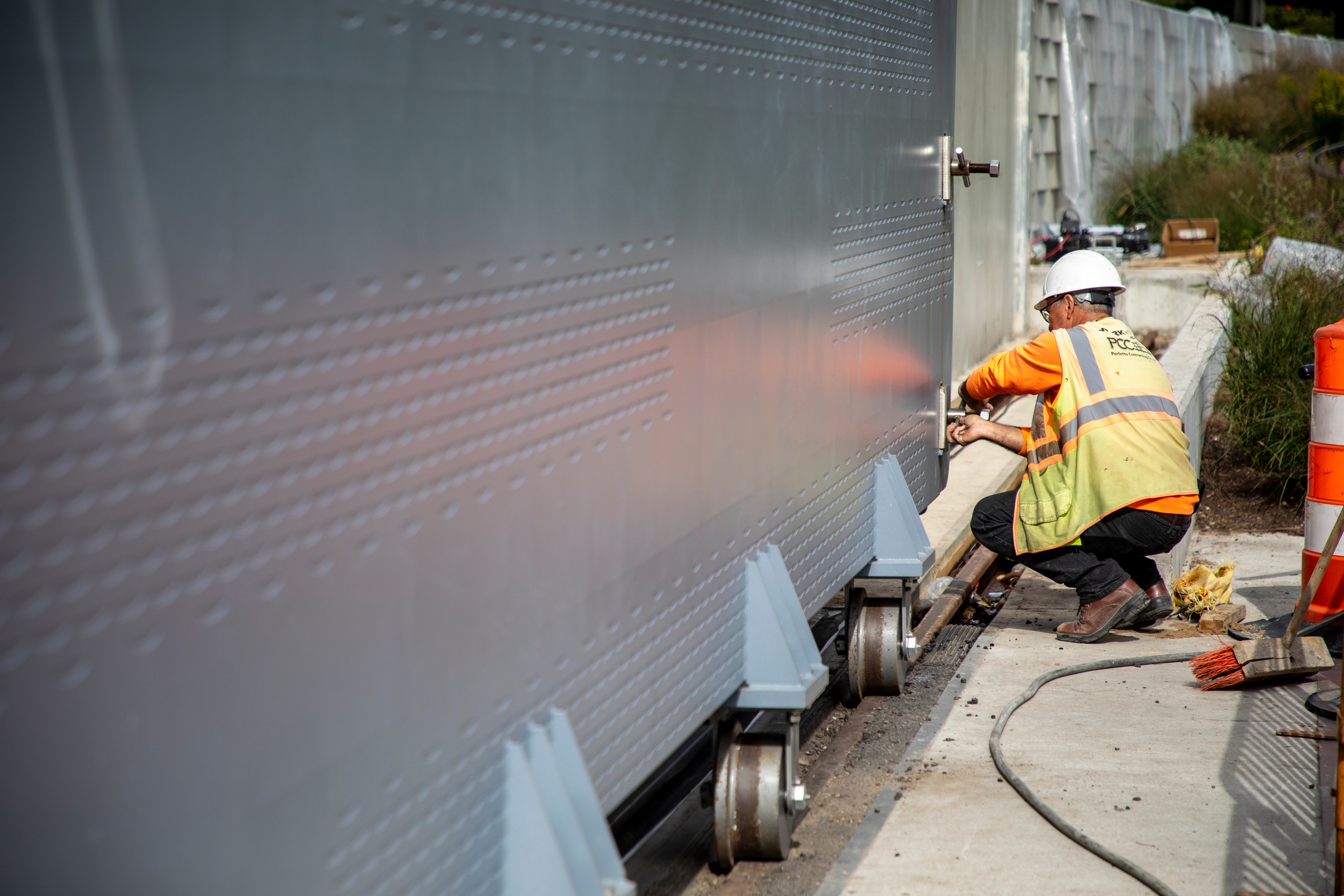 Construction worker in safety gear adjusts a large gray roller floodgate on a track, with tools and traffic cones nearby.