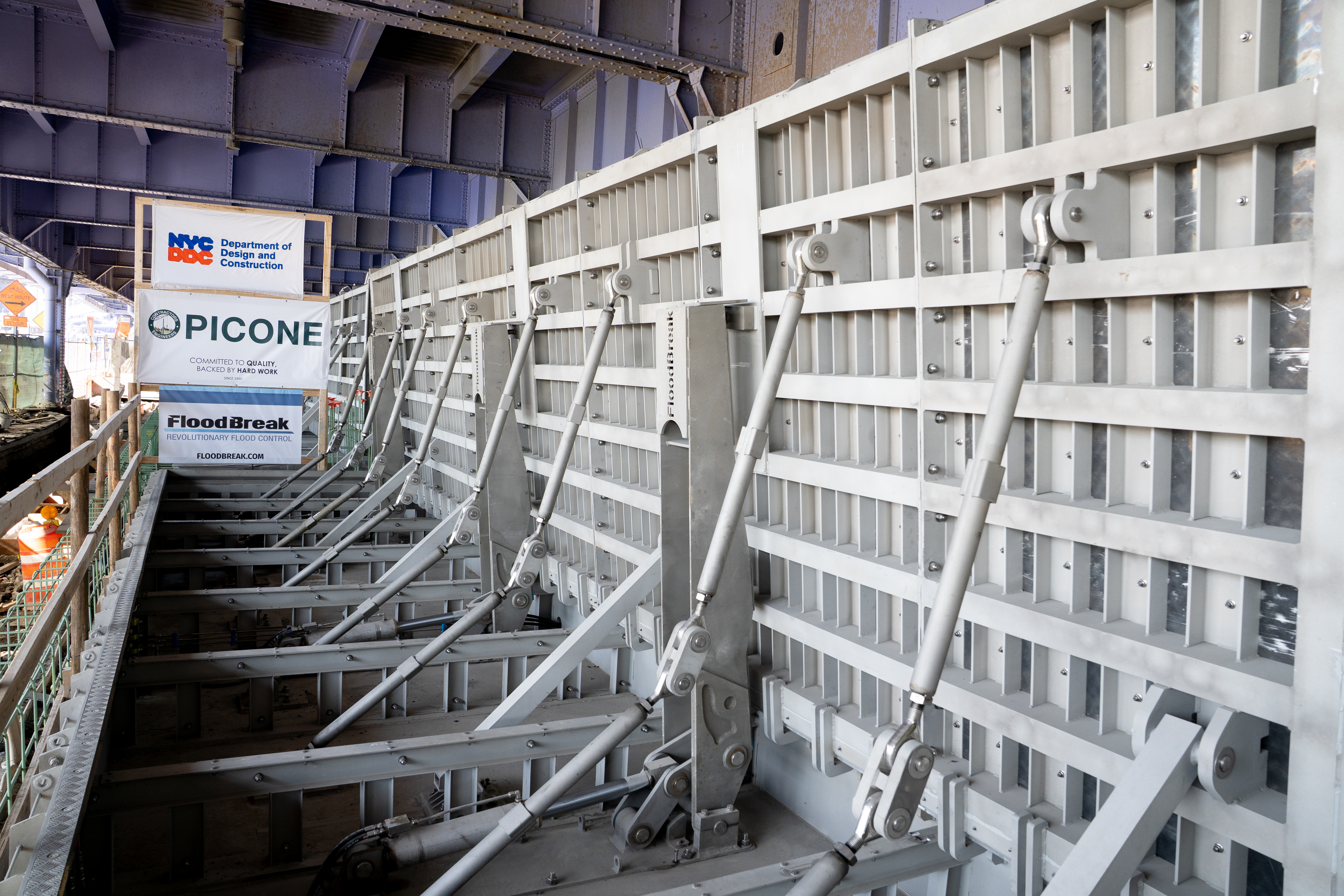 Flip up gate under a bridge, with reinforced metal supports and signage from NYC DDC, Picone, and FloodBreak.