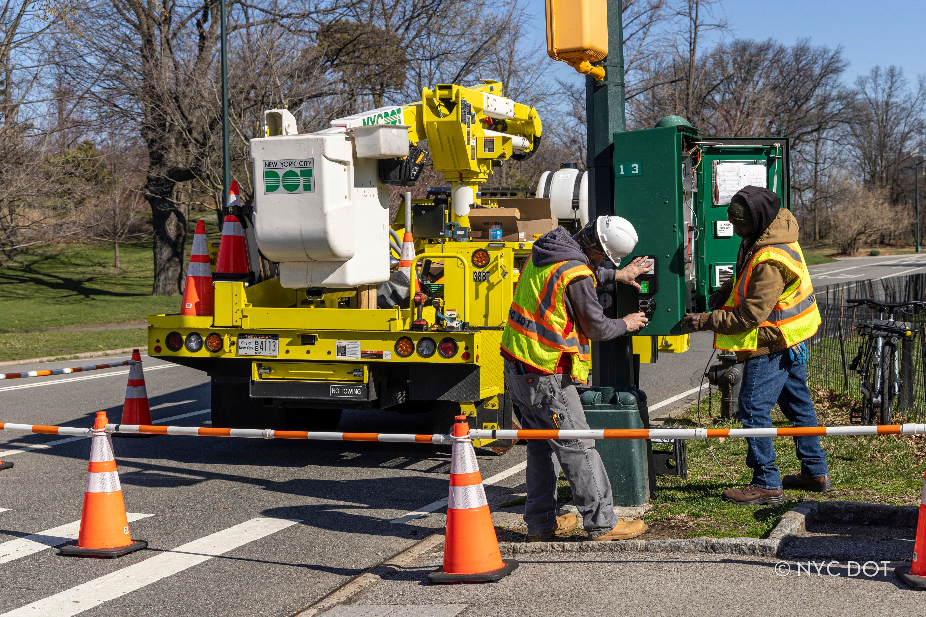 NYC DOT workers in safety gear install a traffic signal control box next to a bucket truck parked near a park.