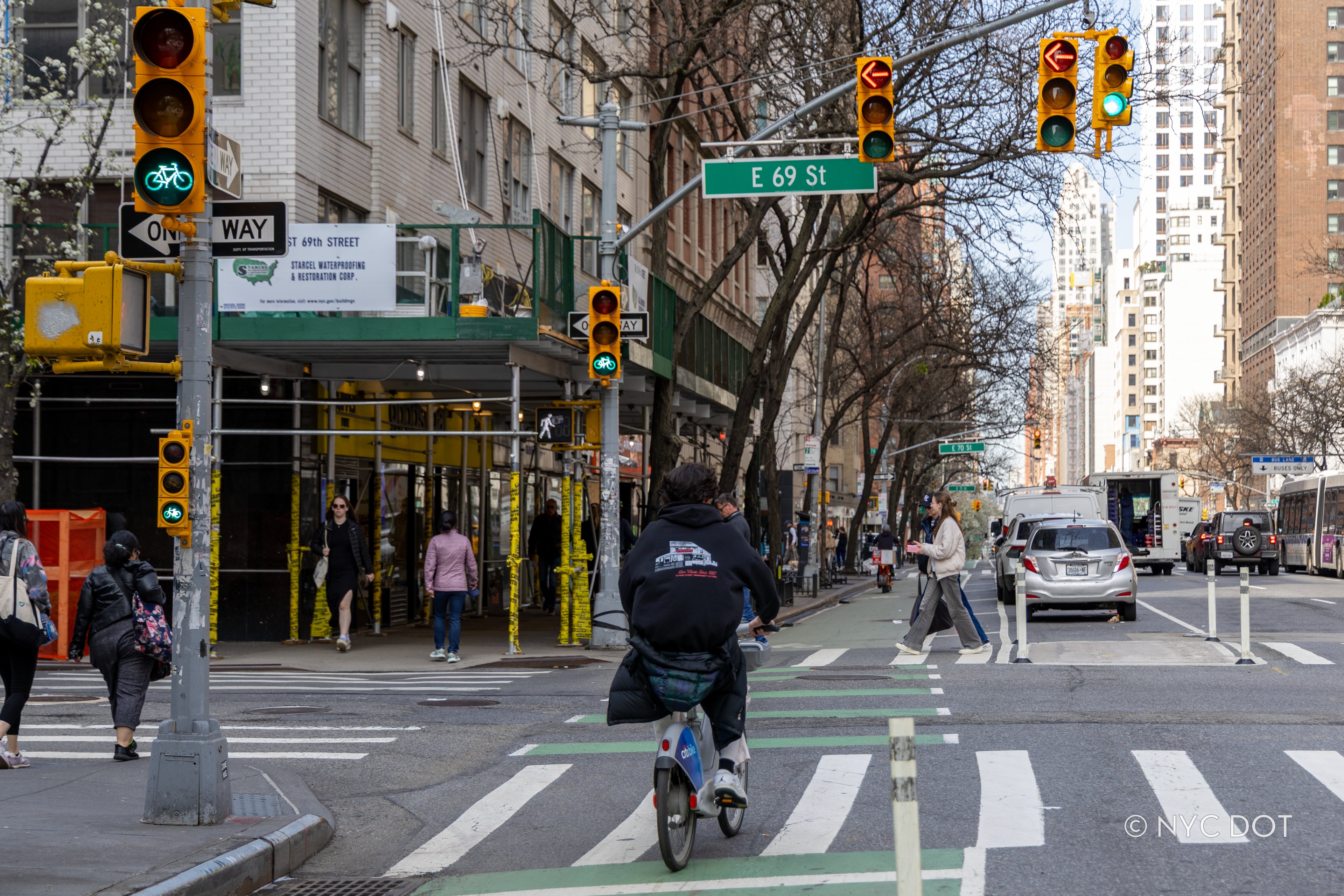 A cyclist rides through a green bike signal at the intersection of E 69th St and 3rd Ave in Manhattan, with pedestrians crossing and traffic lights overhead.
